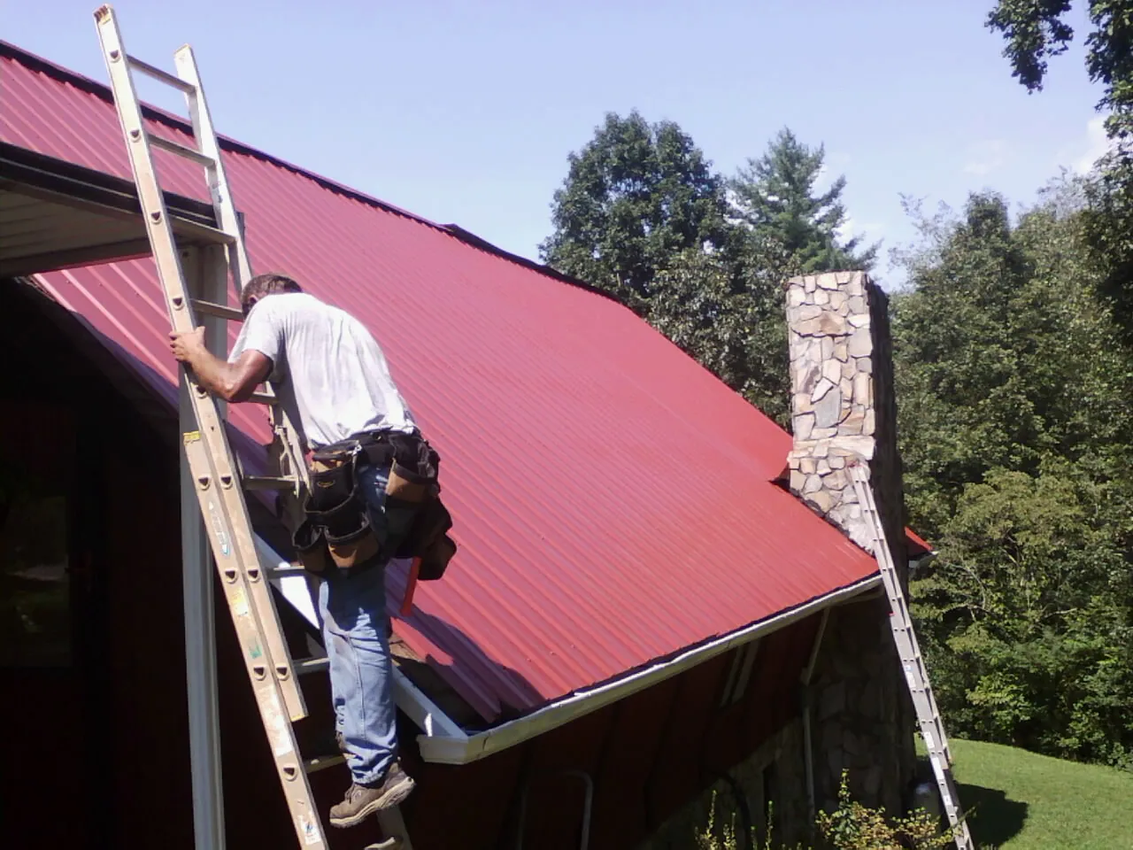 Roofer on a ladder beside a red metal roof and stone chimney — Soffit & Fascia Repair by Roofing Brothers in Huntingdon